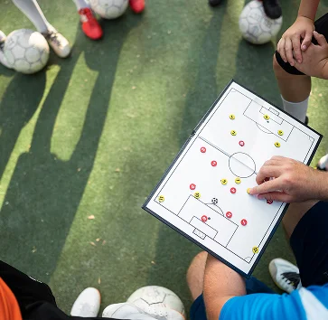 Un entraîneur, muni d'un tableau tactique, explique une stratégie de football à des joueurs rassemblés autour de lui, chacun avec un ballon, sur un terrain ensoleillé.