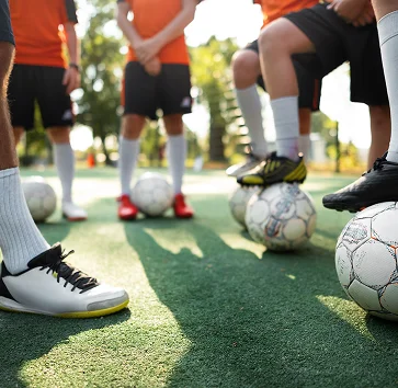 Un groupe de joueurs de football en maillots orange se tient sur un terrain ensoleillé, chacun un pied sur un ballon, symbolisant l'esprit d'équipe et la préparation.