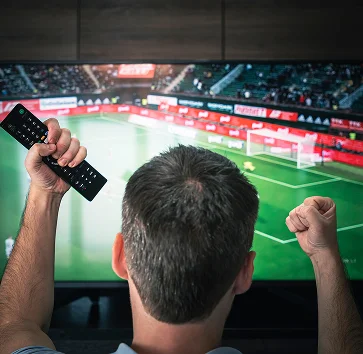 Une personne regarde un match de football à la télévision, poings levés et télécommande à la main, manifestant son enthousiasme et son intérêt pour le jeu.